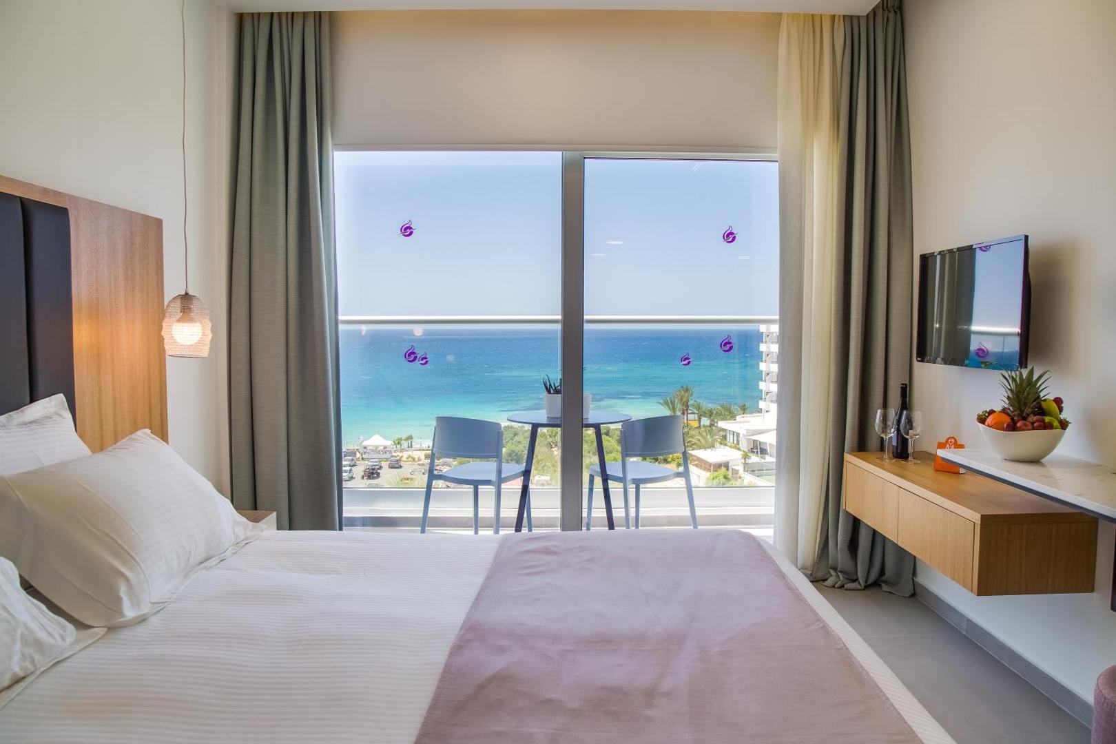 Skats no Napa Mermaid Hotel numura uz Vidusjūru caur lielu logu, gulta priekšplānā un balkons ar galdiņu