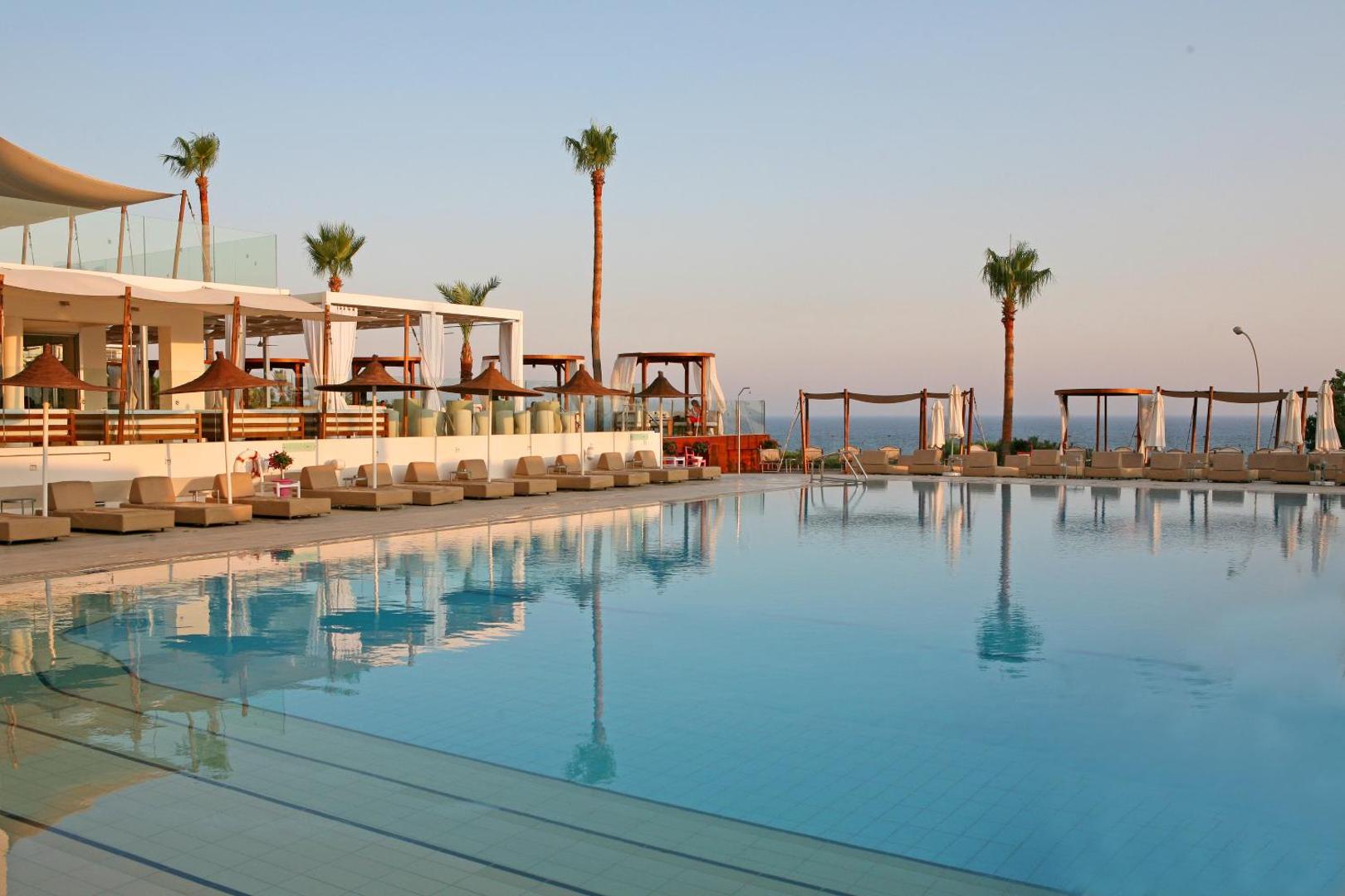 Napa Mermaid Hotel lielais āra baseins ar skatu uz jūru, saulessargiem un atpūtas zonu saulrietā