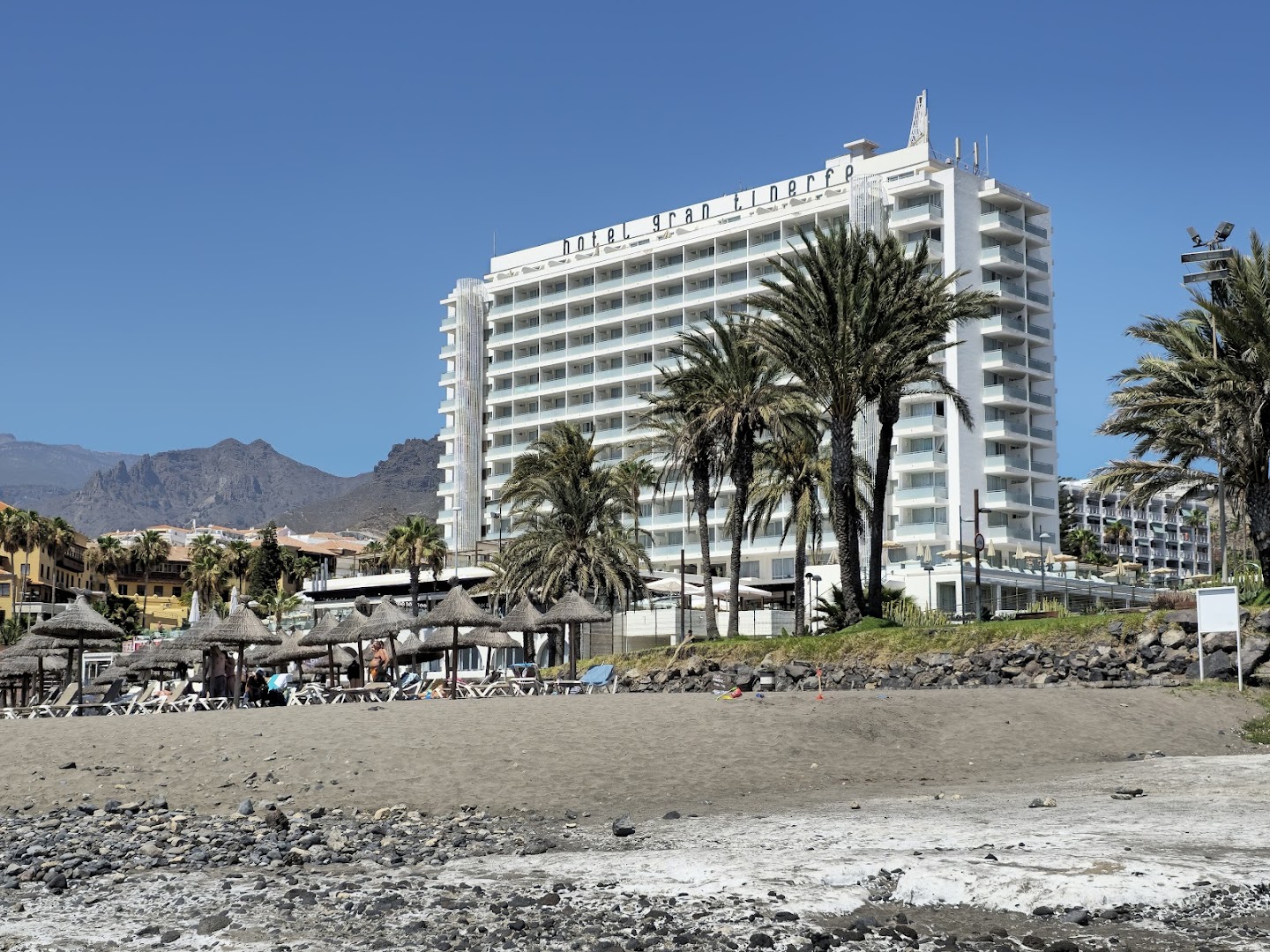 Viesnīcas H10 Gran Tinerfe ēka pie pludmales Tenerifē — gaiša daudzstāvu ēka ar palmām un kalnu fonā