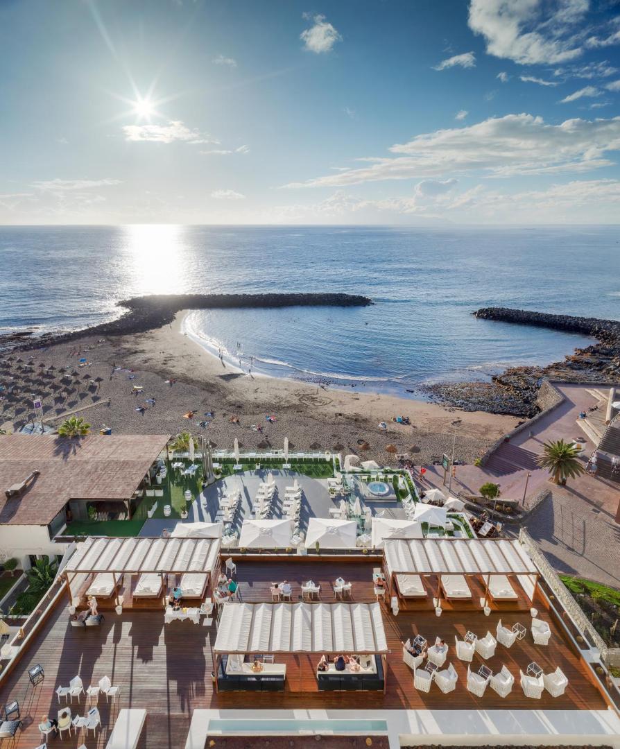 H10 Gran Tinerfe skats no augšas uz viesnīcas terasi, pludmali un okeānu Tenerifē — atpūtas zona ar saulessargiem un dīvāniem