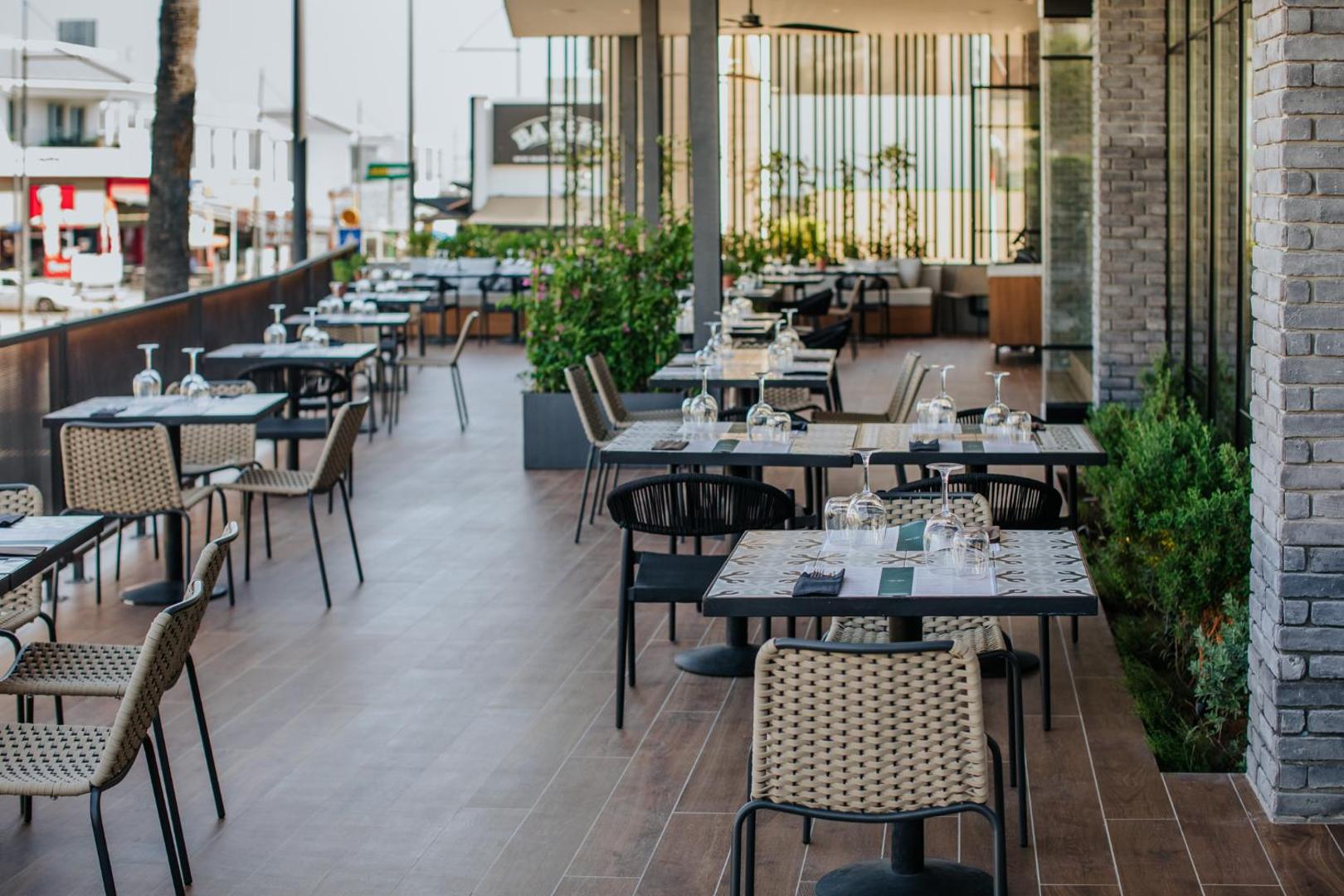 Napa Plaza viesnīcas āra terases restorāns Aijanapā, Kiprā — eleganti klāti galdi ar pītiem krēsliem un pilsētas skatu
