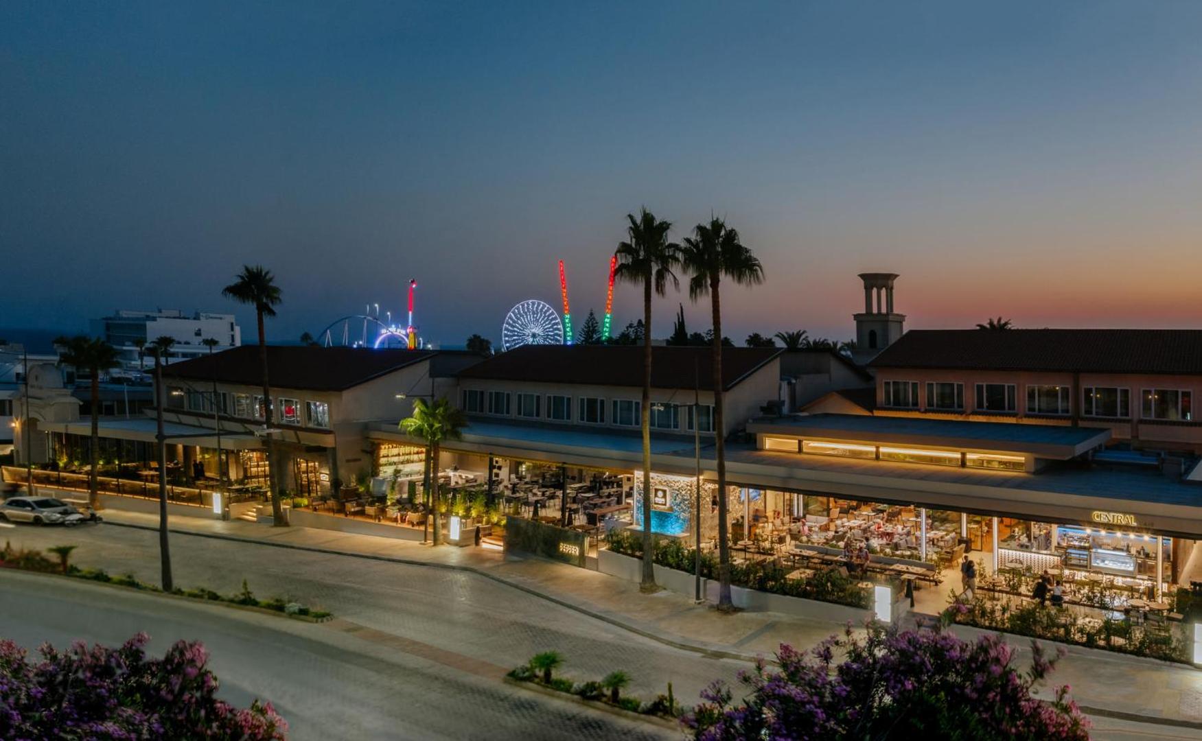 Napa Plaza viesnīcas skats vakarā Ajanapā, Kiprā — izgaismota fasāde, restorāni un panorāmas rats fonā pilsētas centrā