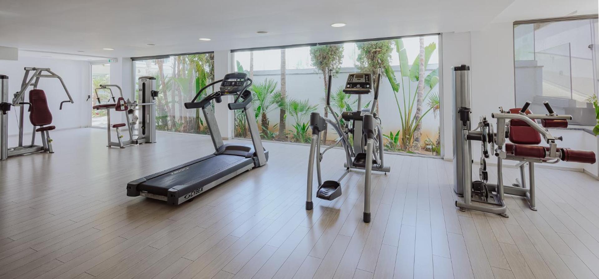 Tasia Maris Oasis modernā fitness zāle ar skatu uz dārzu — skrejceliņi un trenažieri gaišā telpā Aijanapā
