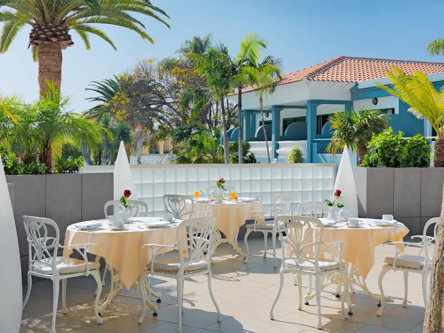Hotel Colon Guanahani restorāna āra terase Tenerifē — balti galdi ar servētām brokastīm palmu un ziedošu krūmu ielenkumā