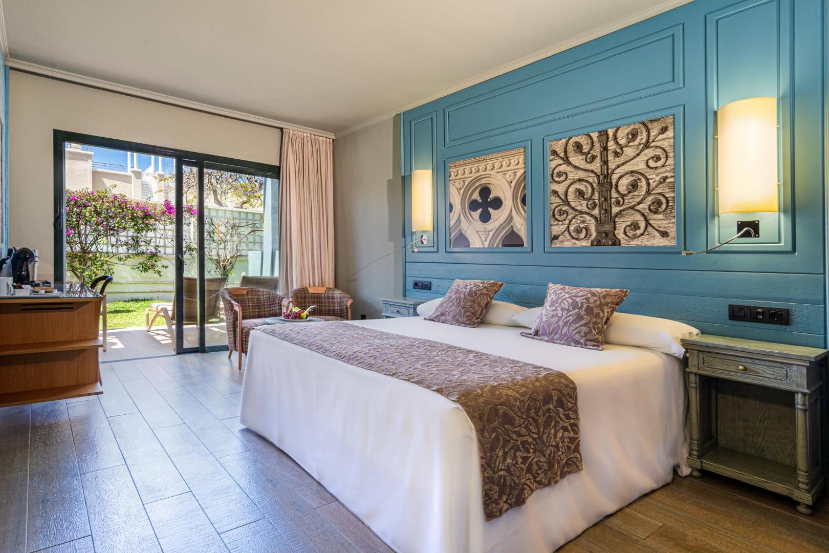 Hotel Colon Guanahani gaišs numurs ar zilu dekoratīvo sienu, lielu gultu un izeju uz dārza terasi Tenerifes piekrastē.