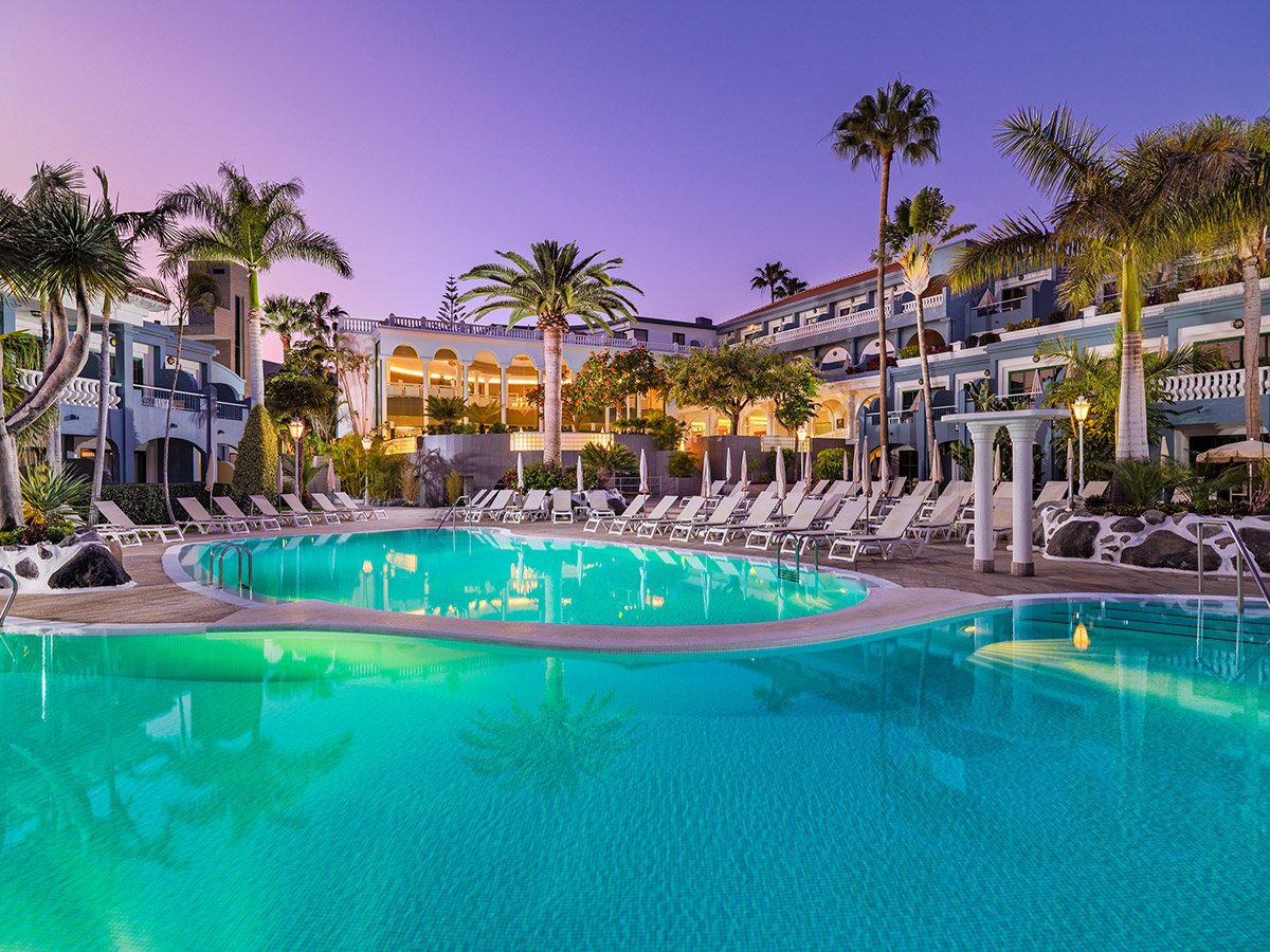 Hotel Colon Guanahani āra baseins vakarā Tenerifē — izgaismota atpūtas zona ar palmām un sauļošanās krēsliem pie baseina saulrietā