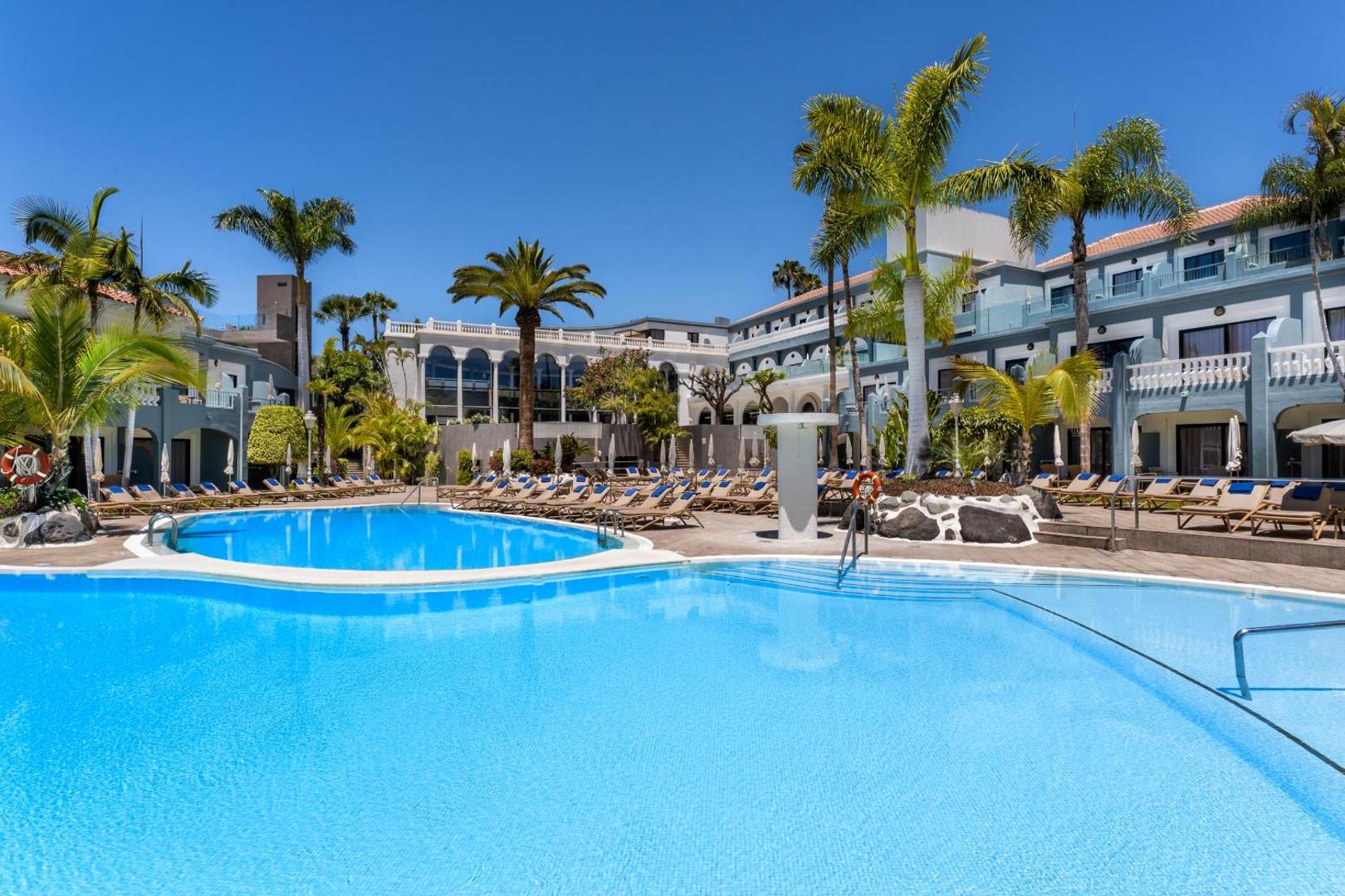 Hotel Colon Guanahani galvenais baseins dienas laikā Tenerifē — plaša sauļošanās zona ar krēsliem un viesnīcas korpuss fonā