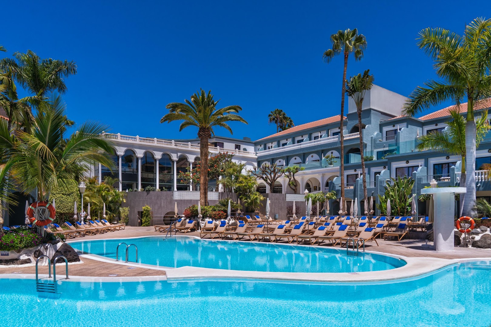 Hotel Colon Guanahani āra baseins ar tirkīza ūdeni, sauļošanās krēsliem un palmām pie viesnīcas ēkas Tenerifē.