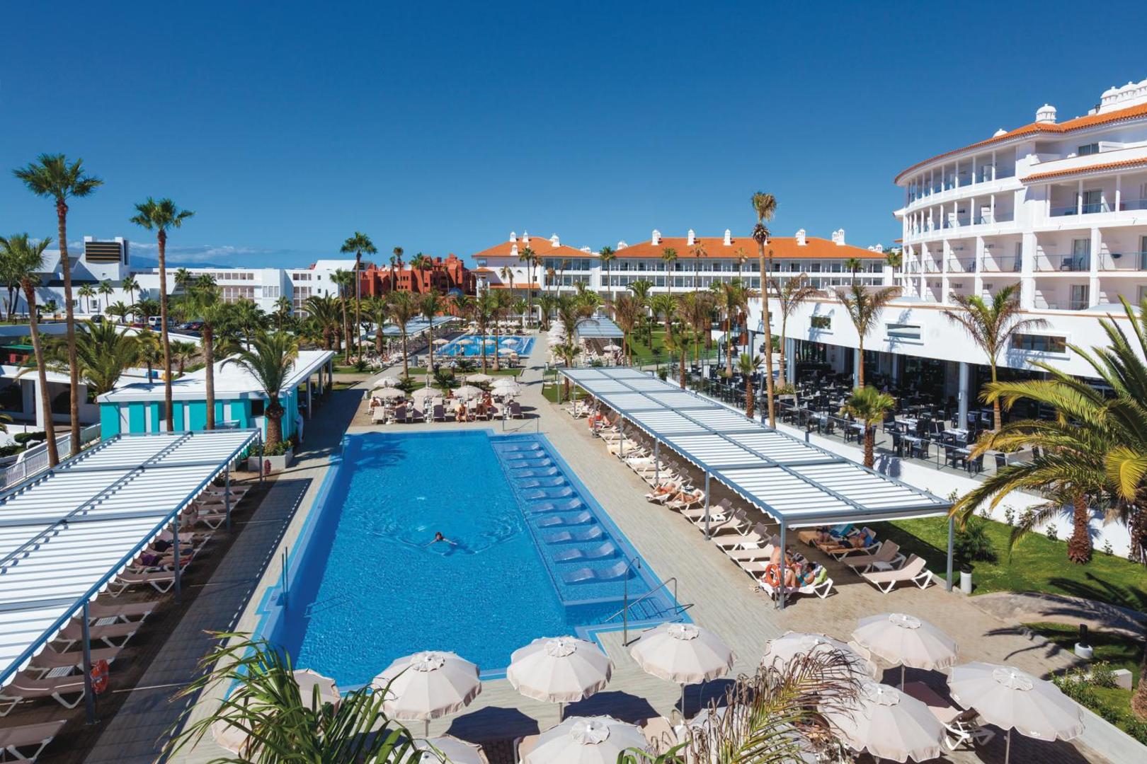 Riu Arecas Hotel āra baseinu zona ar sauļošanās krēsliem, saulessargiem un palmu dārzu Tenerifē — skats uz viesnīcas ēku un terasēm