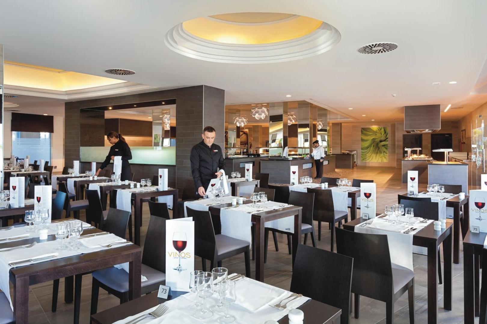 Riu Arecas Hotel restorāna interjers ar tumša koka galdiem un vīna kartēm — profesionāli viesmīļi apkalpo viesus vakariņu laikā