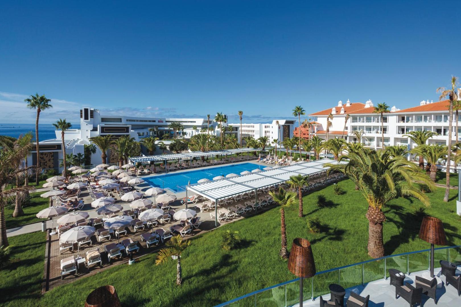 Viesnīcas Riu Arecas āra baseins Tenerifē ar sauļošanās krēsliem, saulessargiem un palmu dārzu piekrastē saulainā dienā.
