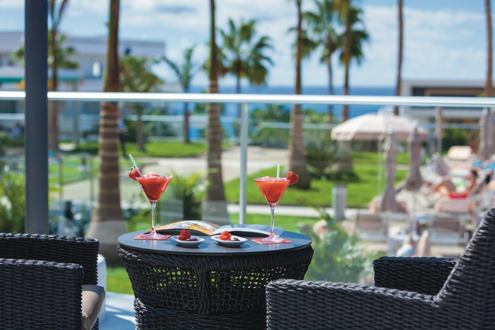 Riu Arecas Hotel terase ar skatu uz palmām un okeānu, uz galda divi sarkani kokteiļi ar zemenēm