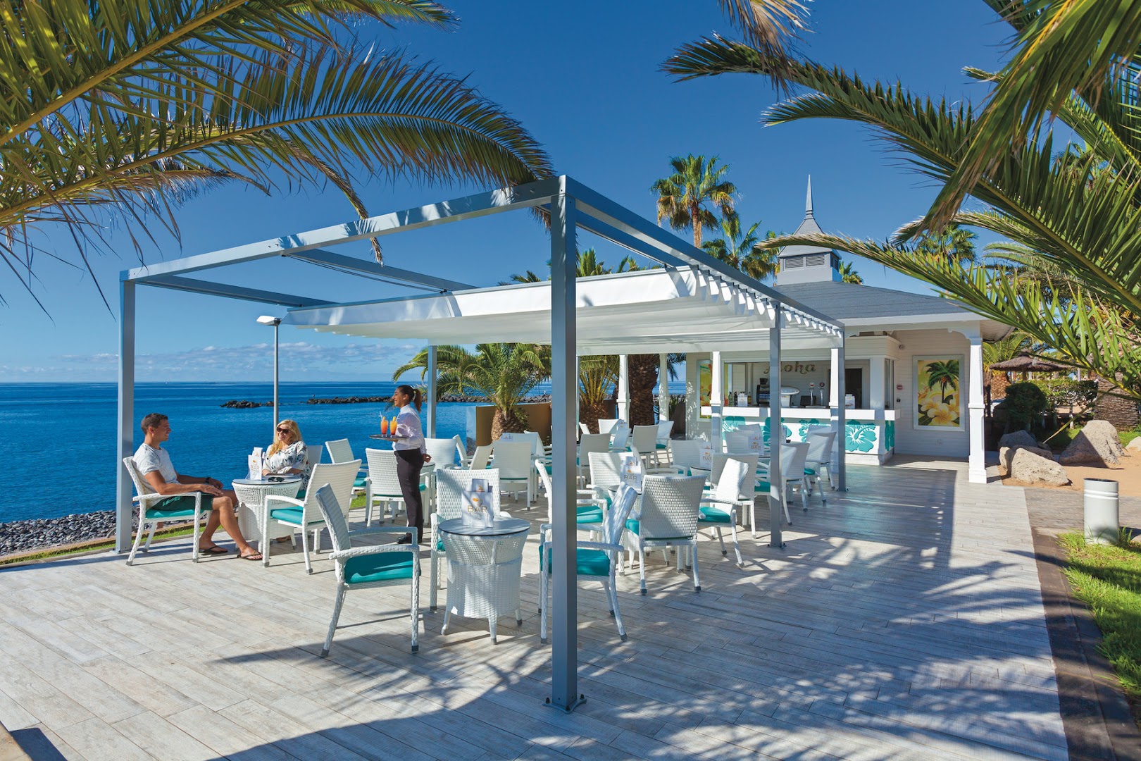 Riu Arecas Hotel āra bārs pie okeāna Tenerifē — gaiša terase ar baltām mēbelēm, saulessargiem un skatu uz zilo jūru
