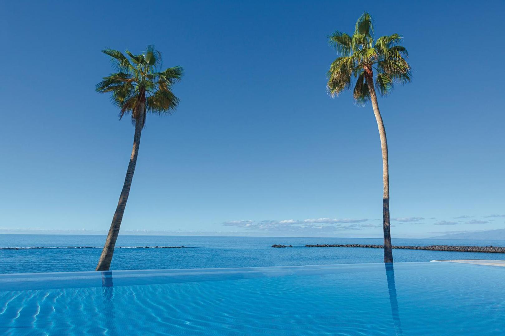 Riu Arecas Hotel bezgalības baseins ar skatu uz zilo okeānu un divām palmām pret skaidrām debesīm Tenerifē
