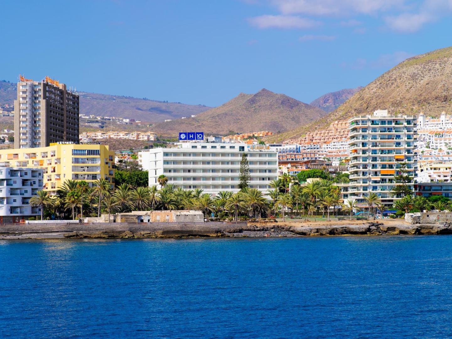 Skats no okeāna uz H10 Big Sur viesnīcu Tenerifes piekrastē — balta ēka uz kalnu un palmu fona