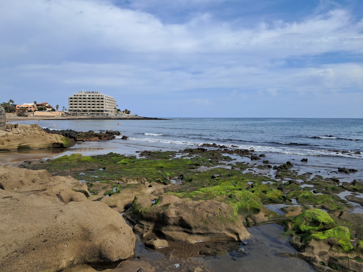 Skats uz KN Arenas del Mar viesnīcas ēku no akmeņainās pludmales Tenerifē — okeāna krasts ar zaļām sūnām un klintīm