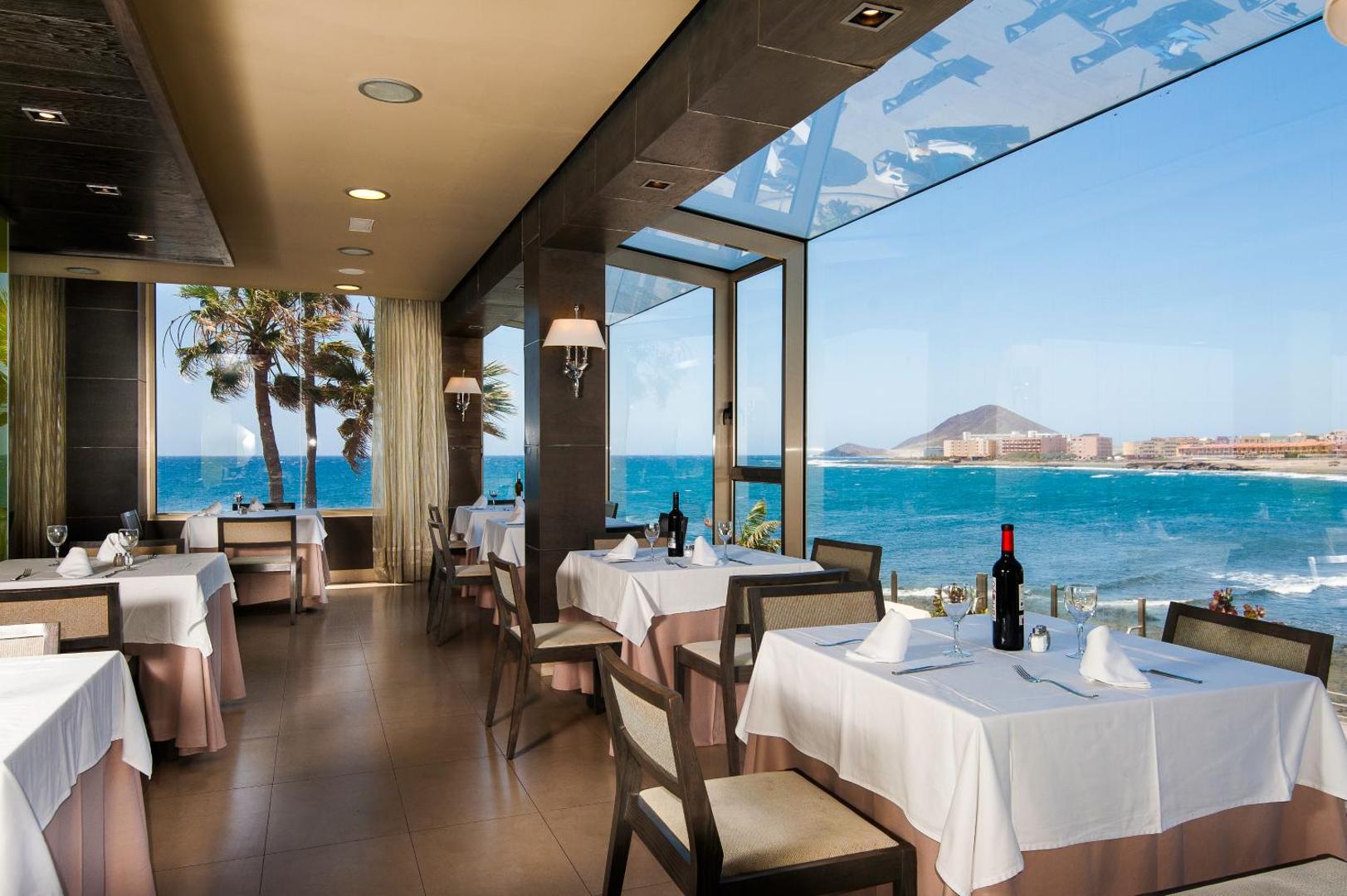 KN Arenas del Mar viesnīcas restorāns ar panorāmas logiem uz okeānu Tenerifē — balti klāti galdi un skats uz piekrasti