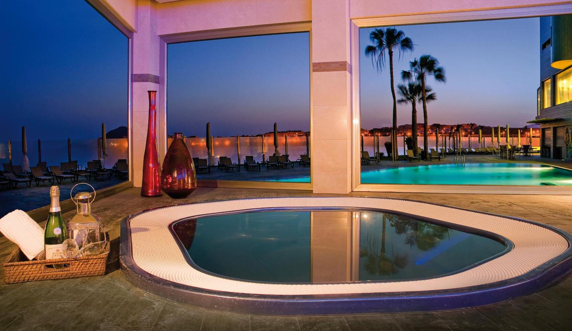 KN Arenas del Mar spa zonas džakuzi ar skatu uz āra baseinu un palmām saulrietā Tenerifē