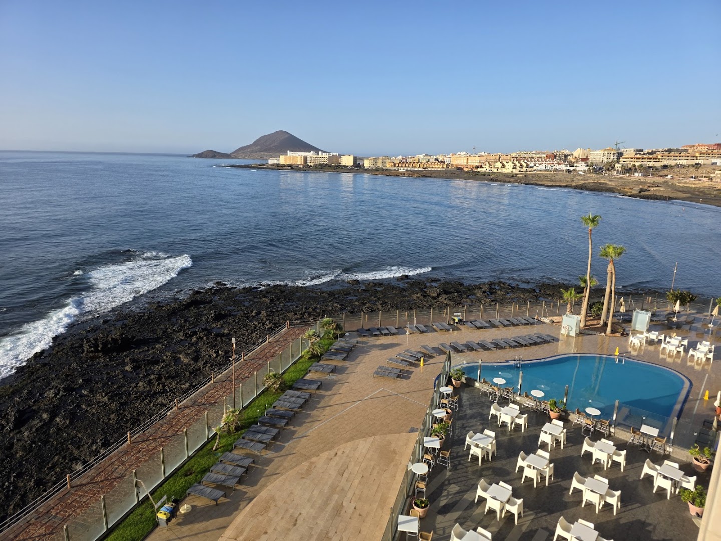 Skats no augšas uz KN Arenas del Mar viesnīcas terasi, āra baseinu, atpūtas krēsliem un akmeņaino Tenerifes piekrasti El Medano