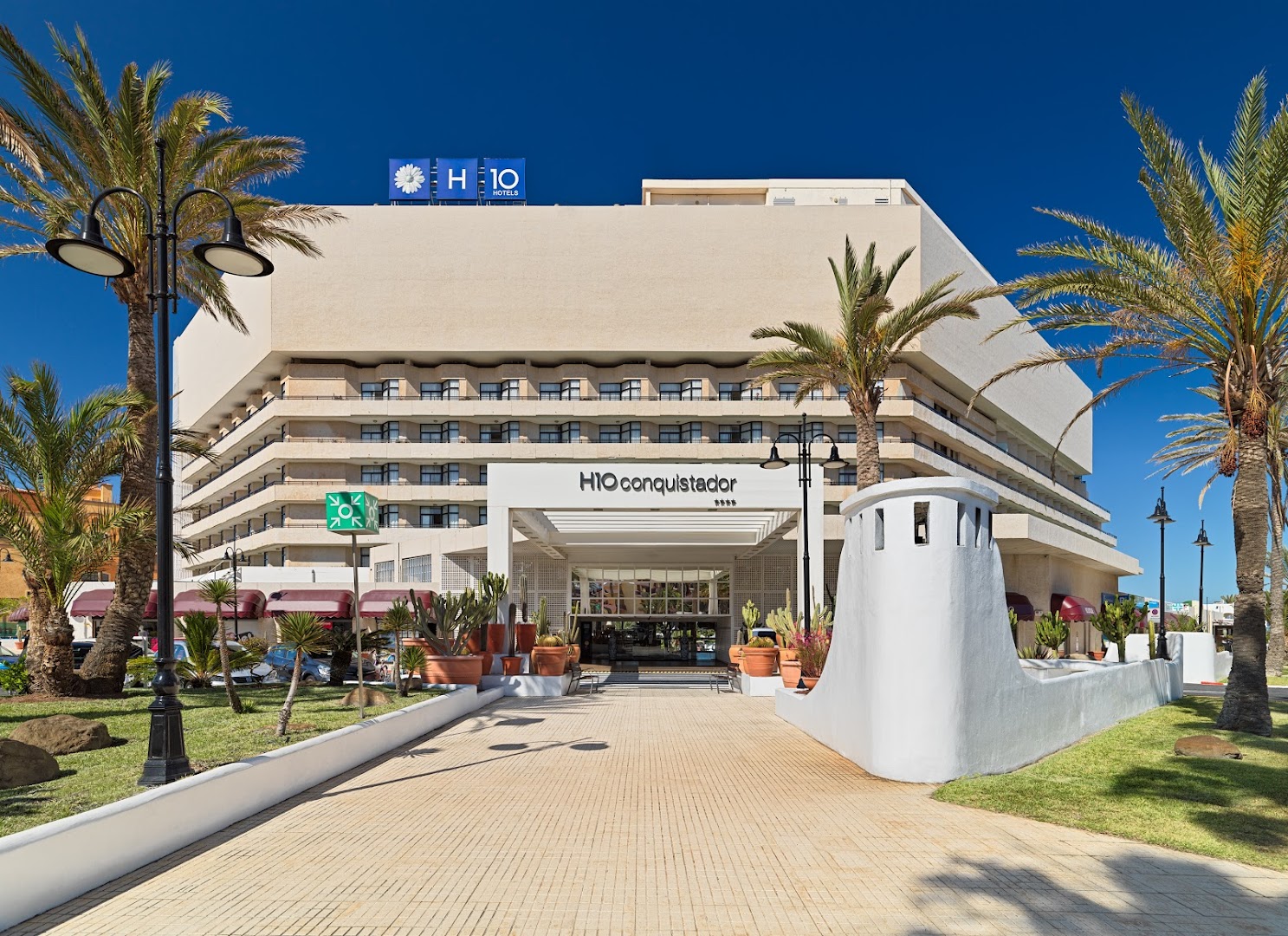 H10 Conquistador viesnīcas ēkas fasāde un galvenā ieeja dienā — palmas, zilas debesis un plaša gājēju zona Tenerifes piekrastē