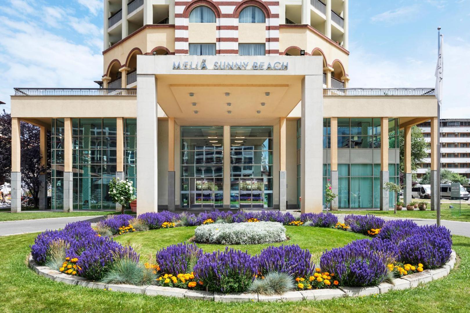Melia Sunny Beach Resort galvenā ieeja ar ziedu dobi priekšplānā un viesnīcas nosaukumu virs stiklotajām durvīm