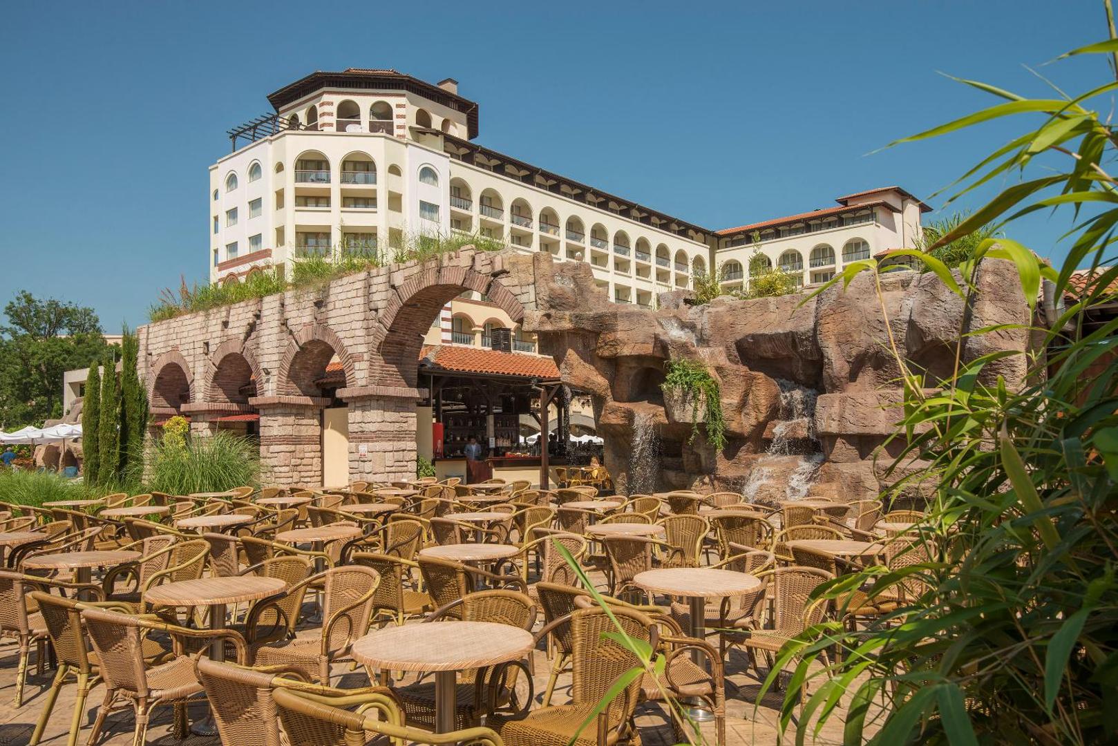 Melia Sunny Beach Resort viesnīcas ēka un āra terase ar pītiem krēsliem, dekoratīvām arkām un mākslīgo ūdenskritumu saulainā dienā