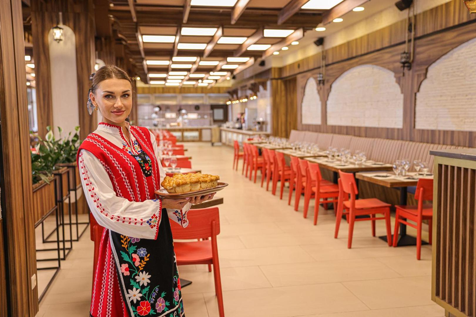 Viesmīle tradicionālā bulgāru tērpā pasniedz maizi Melia Sunny Beach Resort tematiskajā restorānā ar sarkanām mēbelēm un koka interjeru