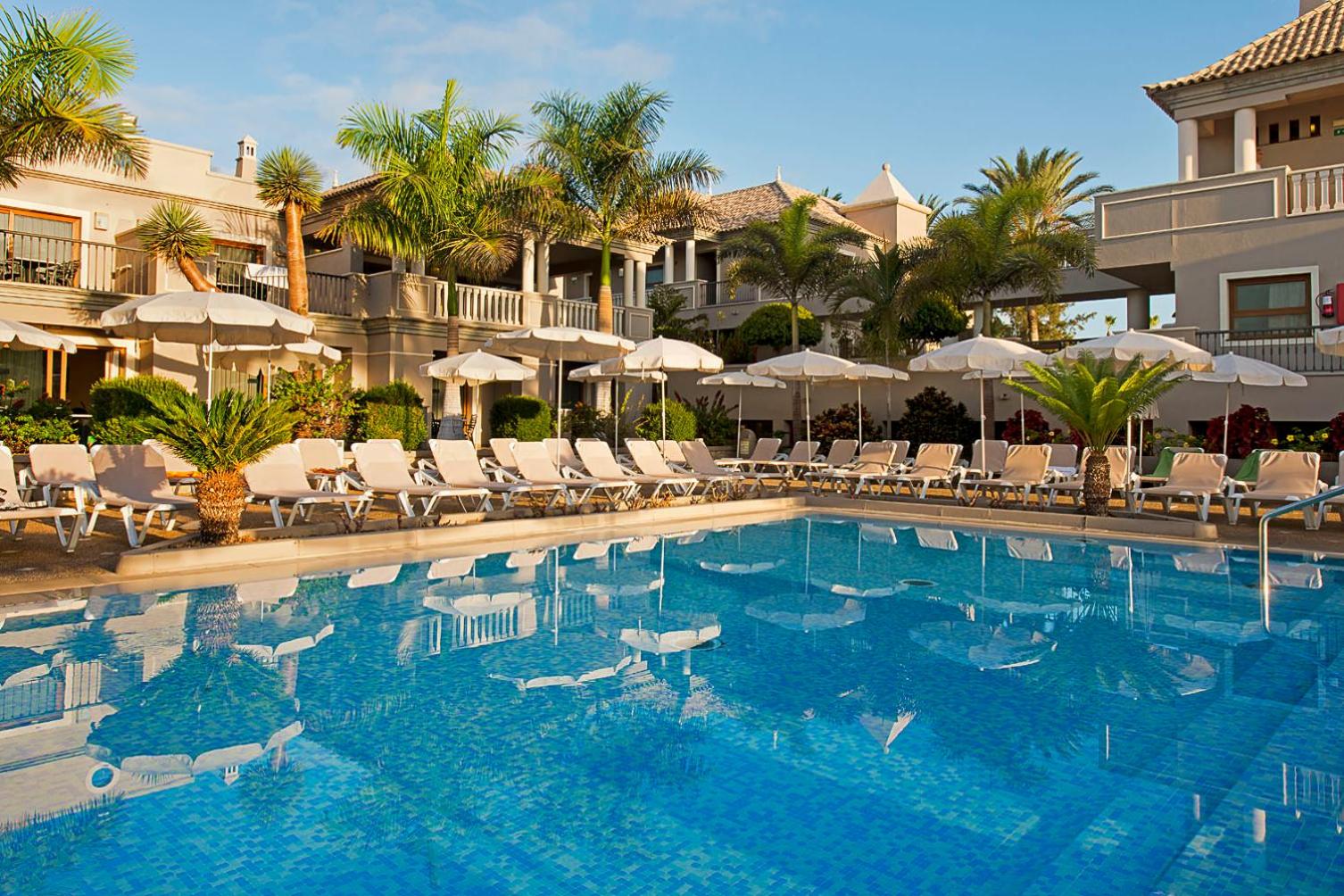 Skats uz Marylanza Suites & Spa zilo baseinu un apkārtējo arhitektūru ar palmām un saulessargiem Tenerifes piekrastē.