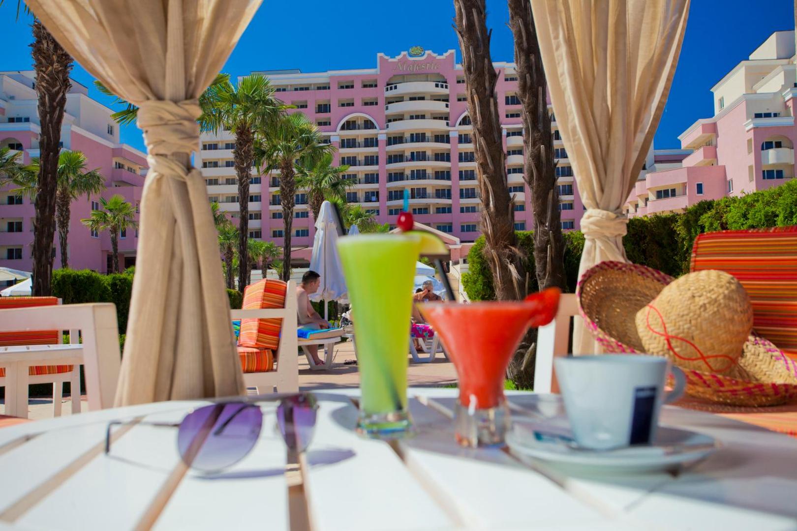 Skats uz DIT Majestic Beach Resort rozā fasādi no terases ar kokteiļiem, saulesbrillēm un palmu ieskautu baseina zonu