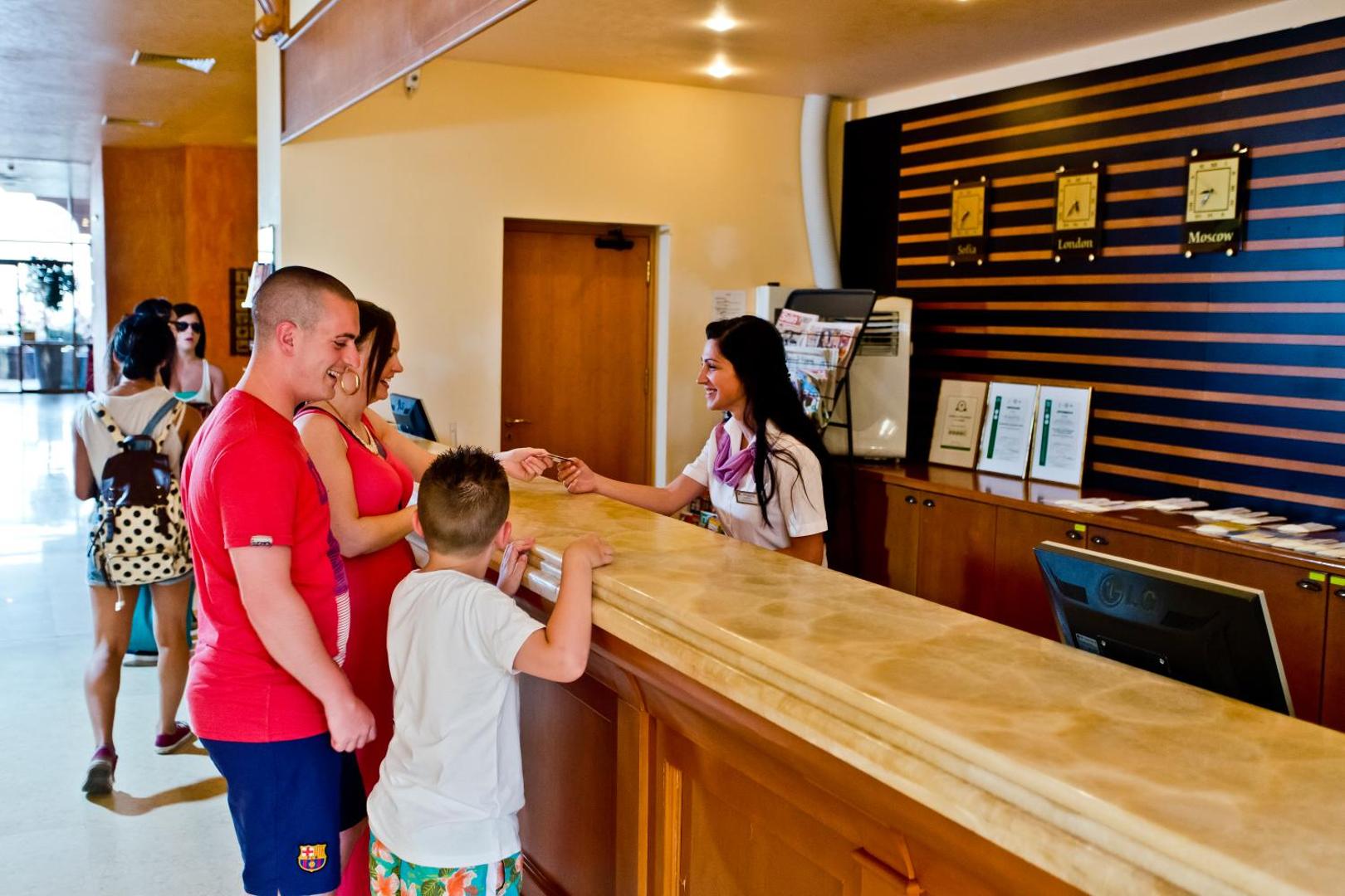 Viesu reģistrācijas process DIT Majestic Beach Resort recepcijā, kur darbiniece apkalpo ģimeni ar bērnu viesnīcas vestibilā