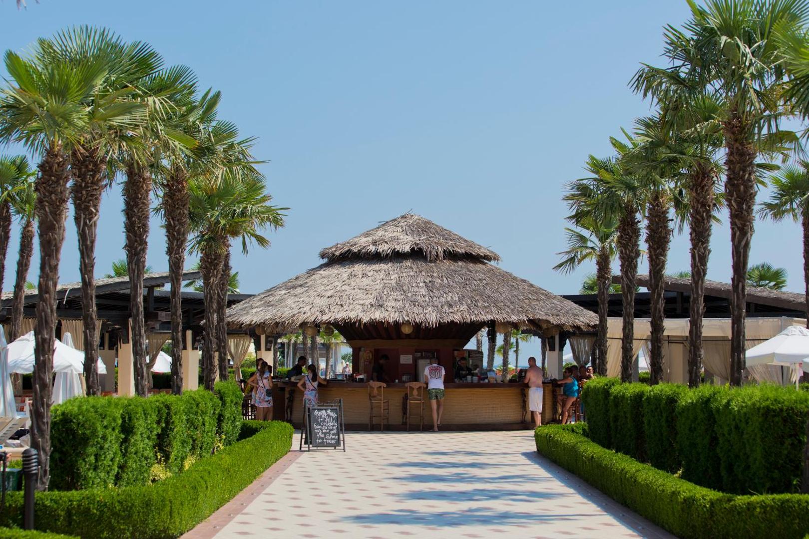 DIT Majestic Beach Resort pludmales bārs ar salmu jumtu — tropiska stila bārs pie baseina, ko ieskauj palmas un zaļumi