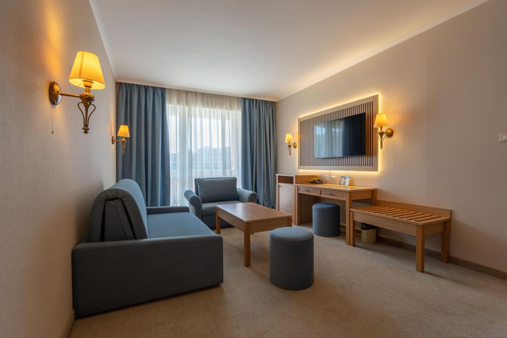 DIT Majestic Beach Resort viesnīcas numura viesistabas zona ar zilu dīvānu, atpūtas krēslu, galdiņu un sienas televizoru