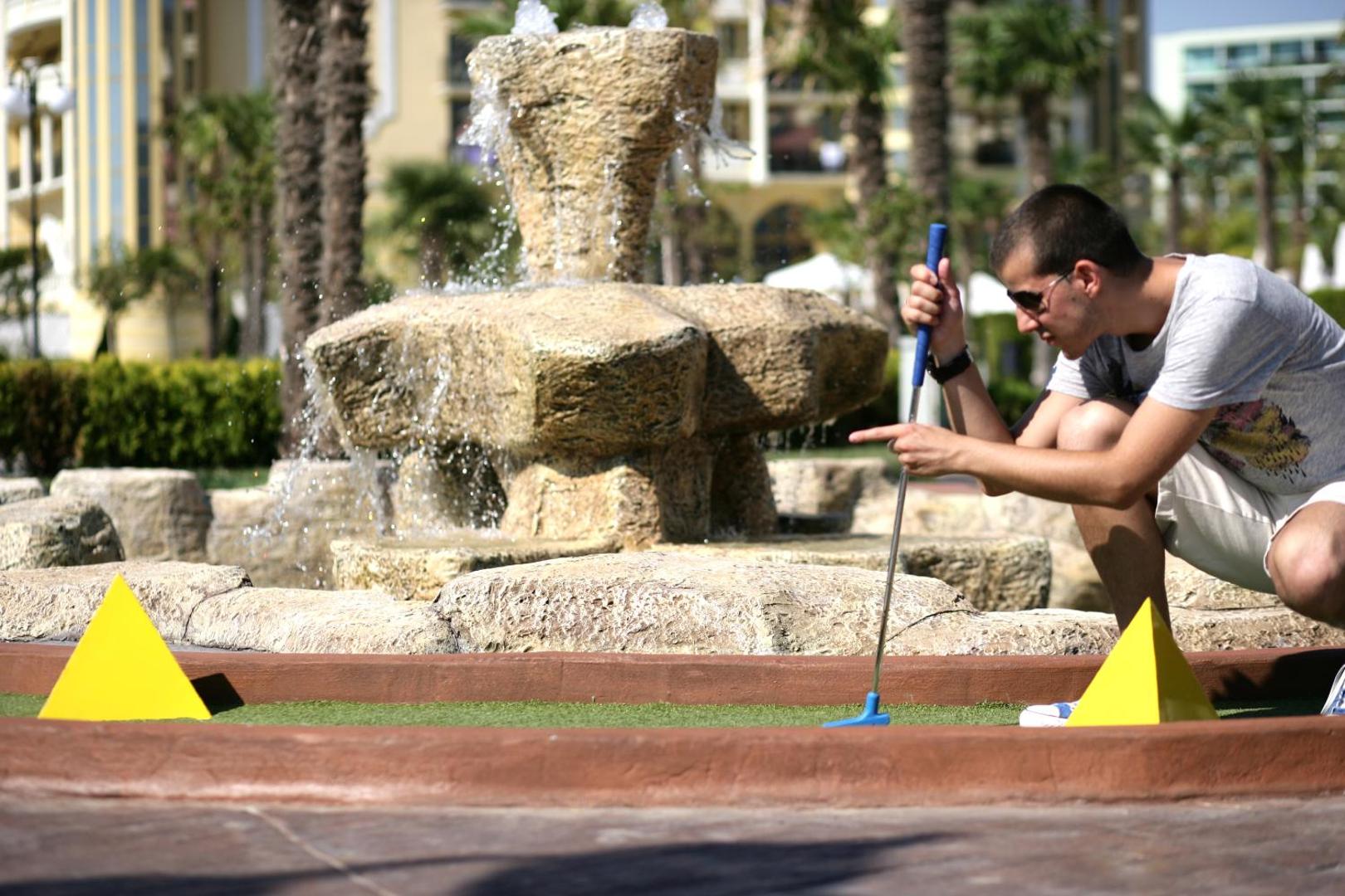 DIT Majestic Beach Resort minigolfa laukums Saulainajā Krastā — viesis spēlē golfu pie akmens strūklakas un palmām