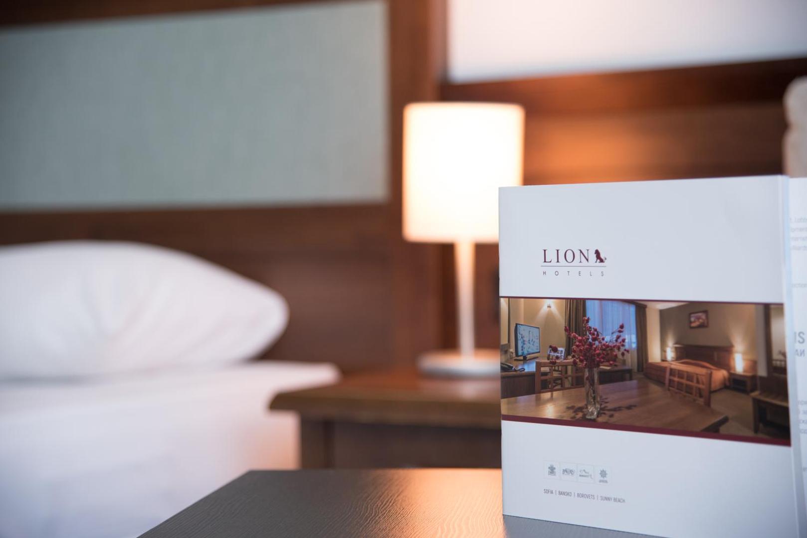 Lion Sunny Beach Hotel informatīvais buklets uz naktsskapīša blakus gultai ar mīkstu apgaismojumu fonā