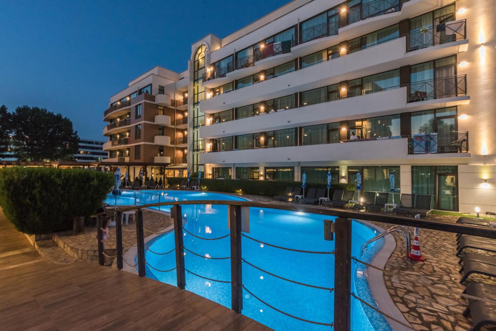 Lion Sunny Beach Hotel fasāde un izgaismots āra baseins vakarā, skats no koka terases Saulainajā krastā