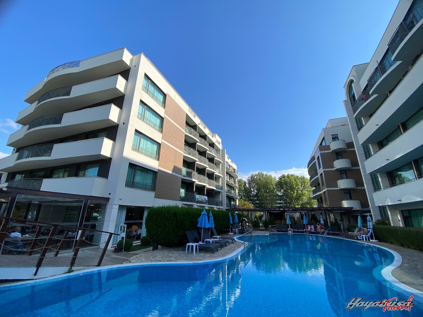 Lion Sunny Beach Hotel modernās korpusa ēkas ap baseinu, zilas debesis un sauļošanās zona Saulainajā krastā