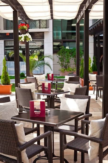 Lion Sunny Beach Hotel āra terase ar pītiem krēsliem un galdiņiem zem nojumes, ko ieskauj zaļumi un ziedi