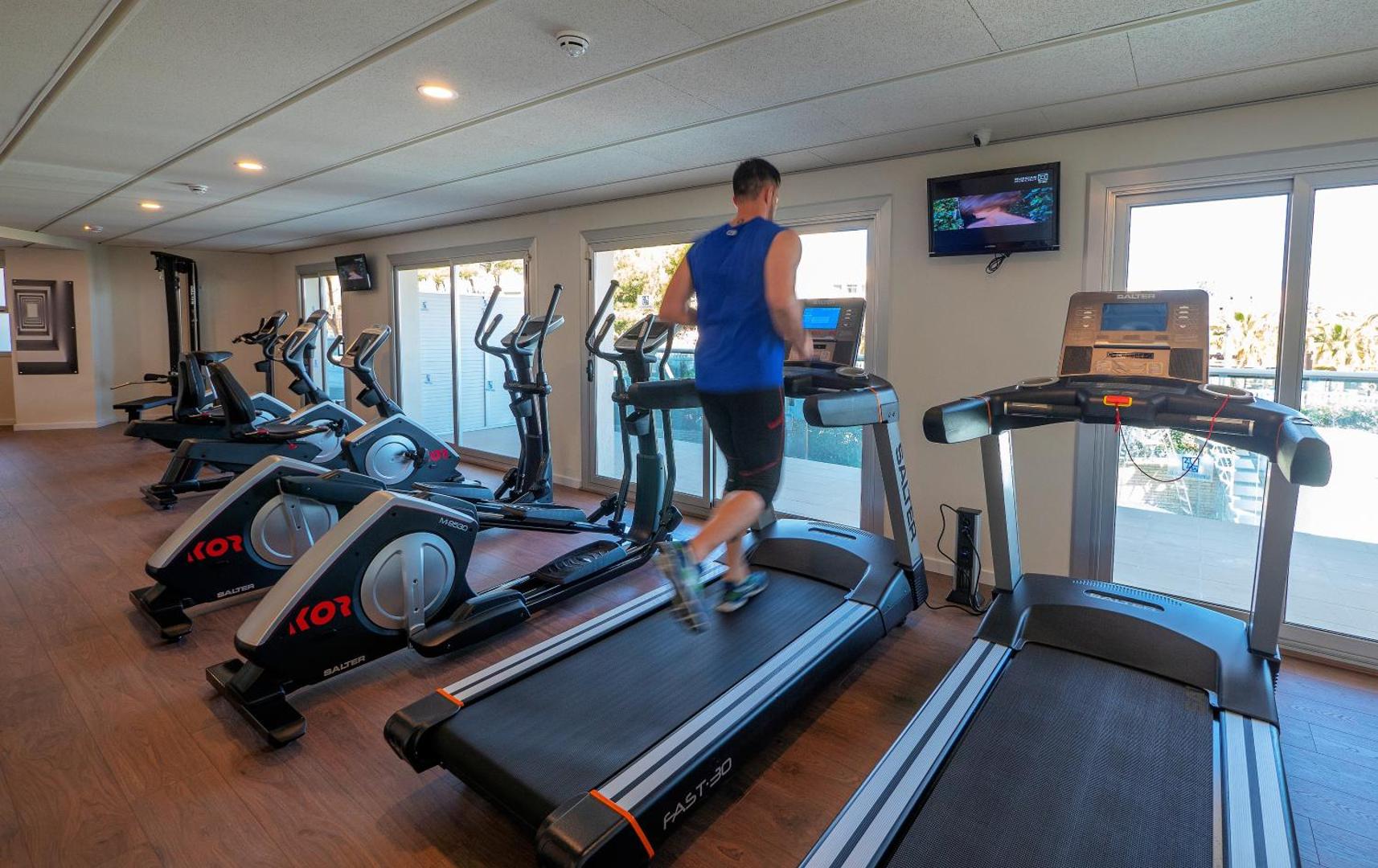Vīrietis sporto Golden Donaire Beach fitnesa centrā uz skrejceliņa — gaiša telpa ar skatu uz āra terasi.