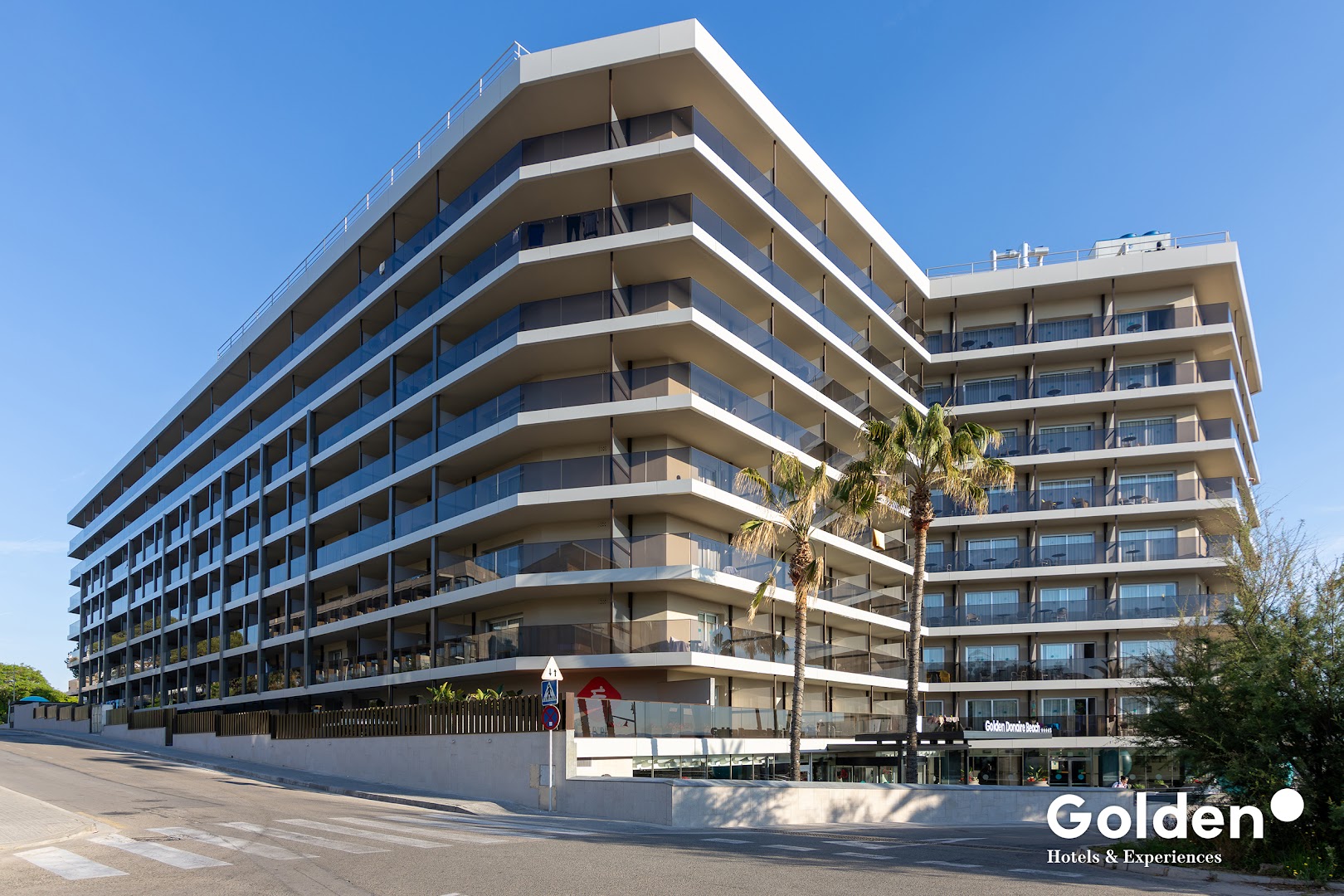 Golden Donaire Beach viesnīcas modernā daudzstāvu ēka ar balkoniem un palmām pie ieejas Kosta Doradā, Spānijā, saulainā dienā.