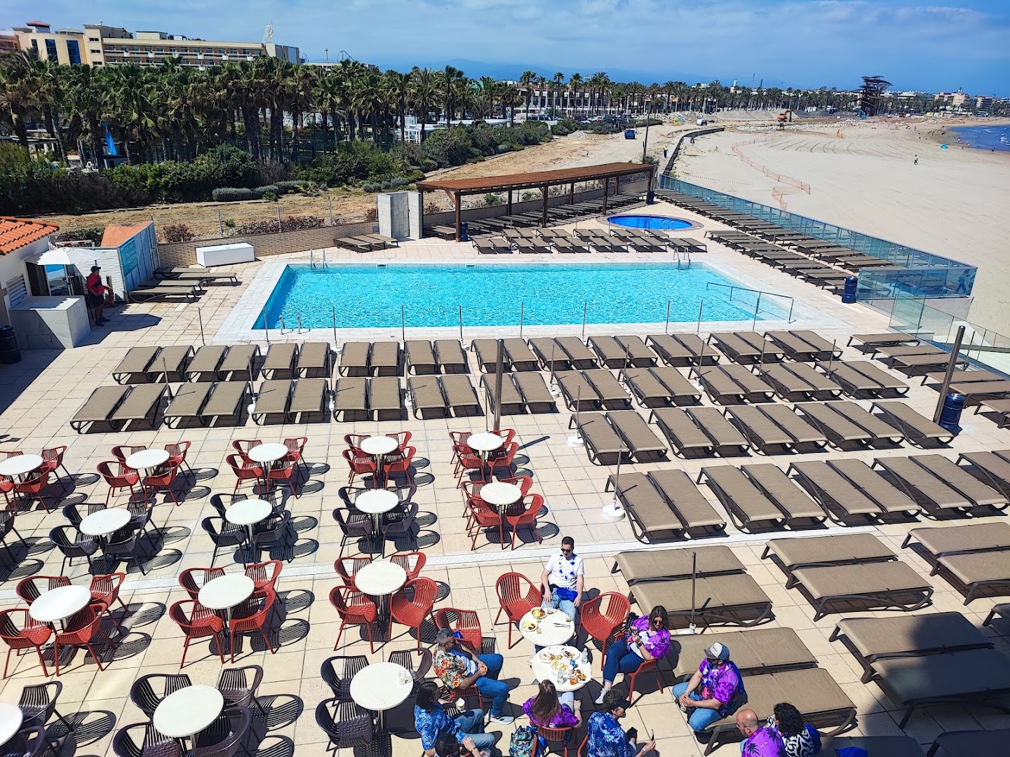 Skats no augšas uz Golden Donaire Beach āra terasi ar kafejnīcas galdiņiem, baseinu un blakus esošo smilšu pludmali