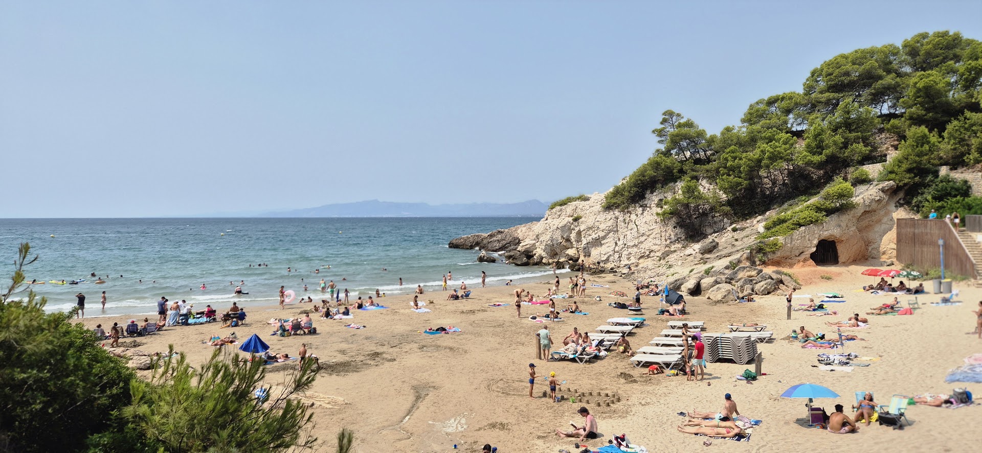 Skats uz smilšu pludmali pie viesnīcas Cala Font Salovā — tirkīzzils ūdens, klintis un atpūtnieki piejūras līcī.