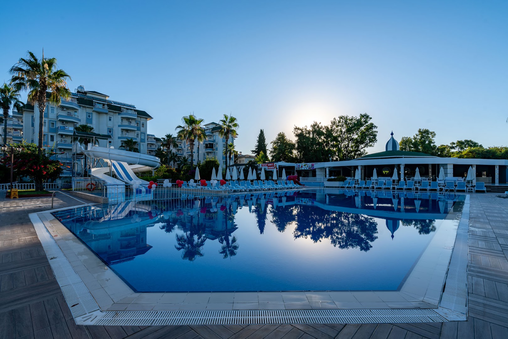 The Garden Beach Hotel āra baseins ar ūdens slidkalniņu un saulessargiem dienas gaismā — skats uz viesnīcas numuru korpusu