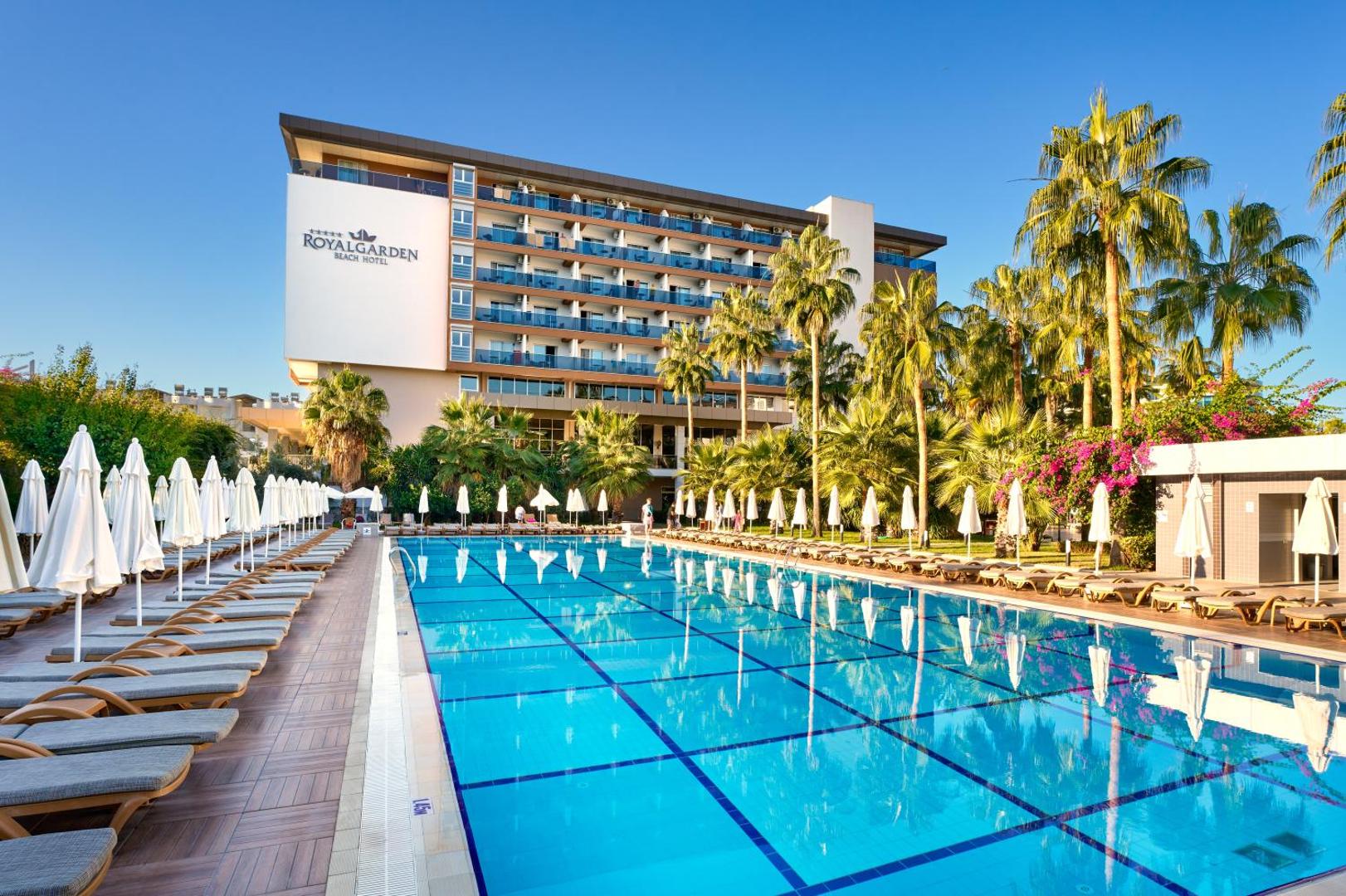The Garden Beach Hotel galvenais baseins ar sauļošanās krēsliem, saulessargiem un viesnīcas ēku fonā Alanjā
