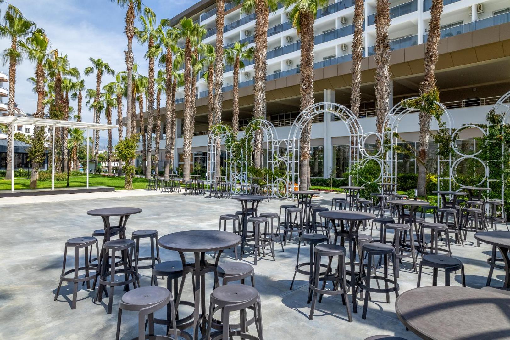 The Garden Beach Hotel āra terase ar bāra galdiem un krēsliem, ko ieskauj augstas palmas un viesnīcas korpuss