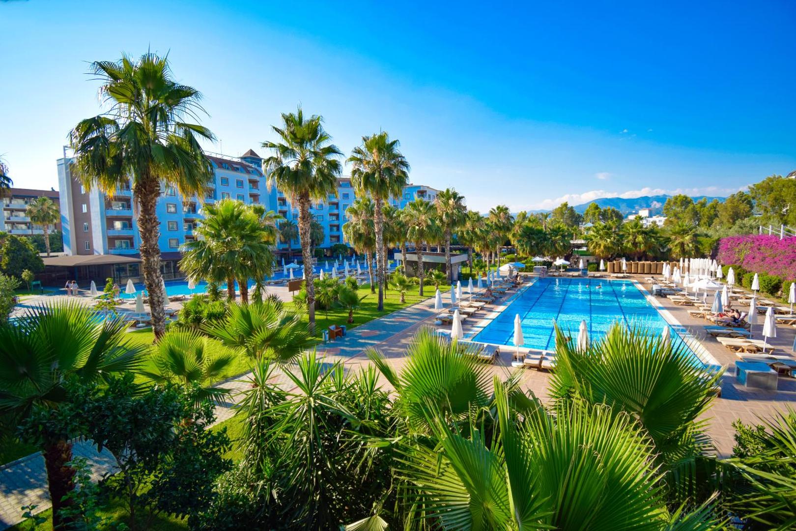 The Garden Beach Hotel plašā teritorija ar āra baseiniem un palmām Alanjas reģionā — skats uz viesnīcas korpusiem un atpūtas zonu