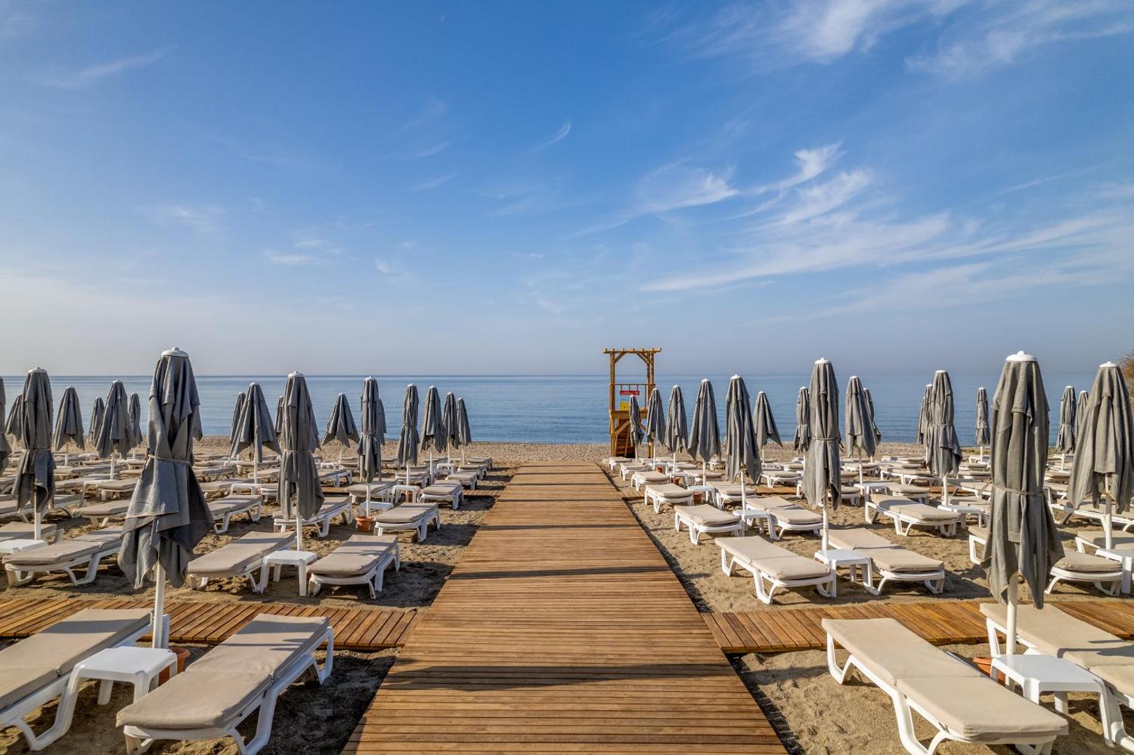 Koka laipa The Garden Beach Hotel pludmalē starp saulessargu rindām ved uz Vidusjūras krastu saulainā dienā