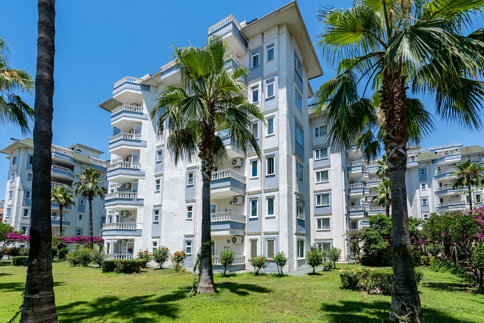 The Garden Beach Hotel daudzstāvu korpuss ar balkoniem un koptu zālāju, ko ieskauj augstas palmas un dekoratīvie krūmi