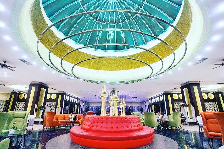 Belconti Resort Hotel lobby halle ar krāsainiem dīvāniem un iespaidīgu stikla kupolu — moderna atpūtas zona ar augstiem griestiem