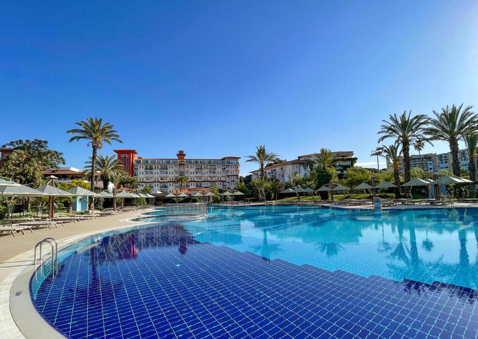 Belconti Resort Hotel lielais peldbaseins ar zilu flīžu apdari, saulessargiem un viesnīcas ēku fonā zem skaidrām debesīm