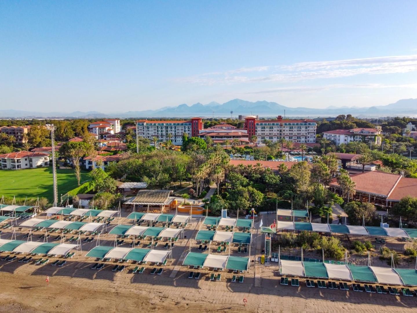 Aerofoto uz Belconti Resort Hotel kompleksu Belekā — galvenā ēka, dārza numuri un pludmales zona uz kalnu fona